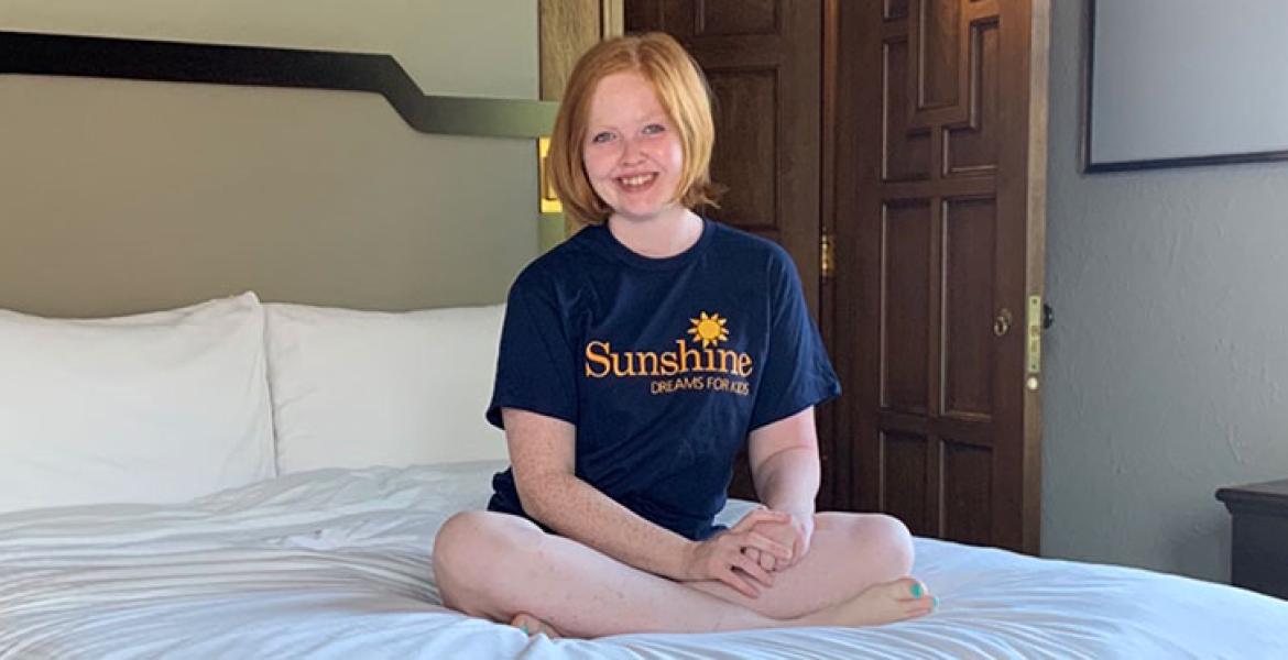 Young woman with fair skin, orange bobbed hair sits cross-legged on king size bed
