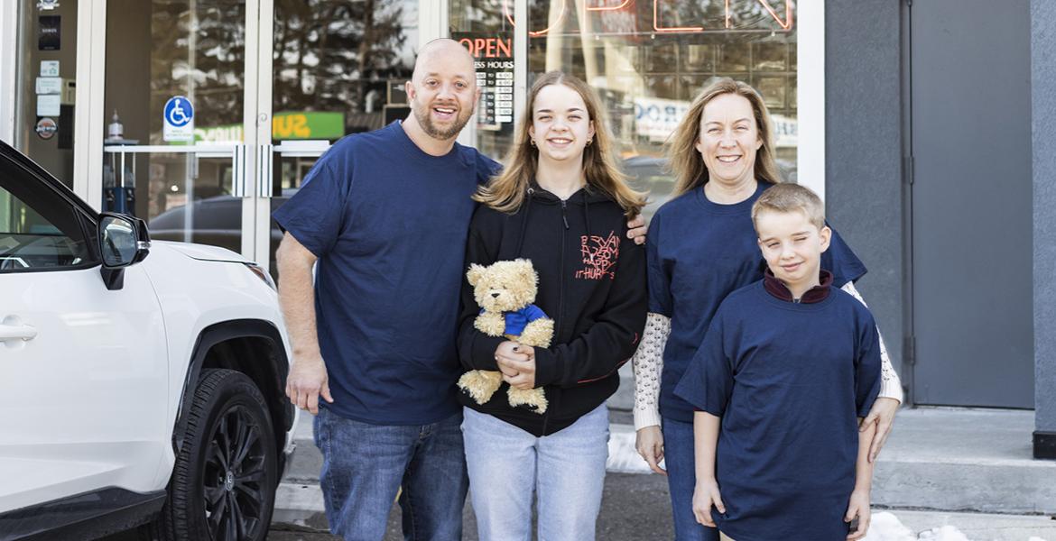 Jocelyne stands in black sweater outside a store with her dad holding her shoulder and her mom and bother to her other side