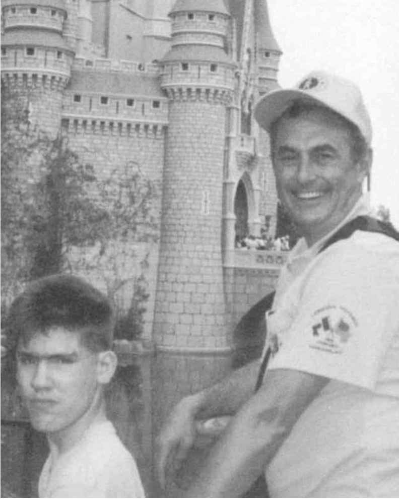 "Man with a big smile wearing Sunshine hat and shirt pushes a teenager in a wheelchair. Photo taken in front of Disney castle."