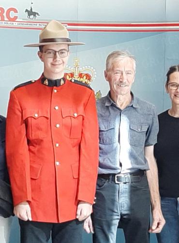 Erik stands in front of RCMP wall in uniform and hat, his parents on each side and two RCMP officers on each end