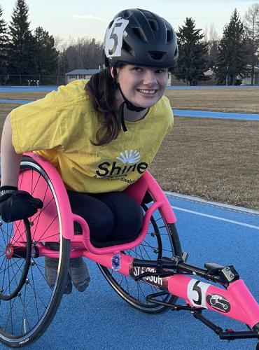 Maggie is seated in her hot pink racing wheelchair on a bright blue outdoor track wearing a yellow Shine tshirt and black helmet