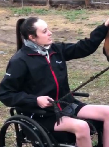 Sabrina seated in a wheelchair, pets the nose of her horse while holding his reins