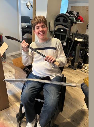 Blake seated in wheelchair holding sledge hockey sticks beside a large box