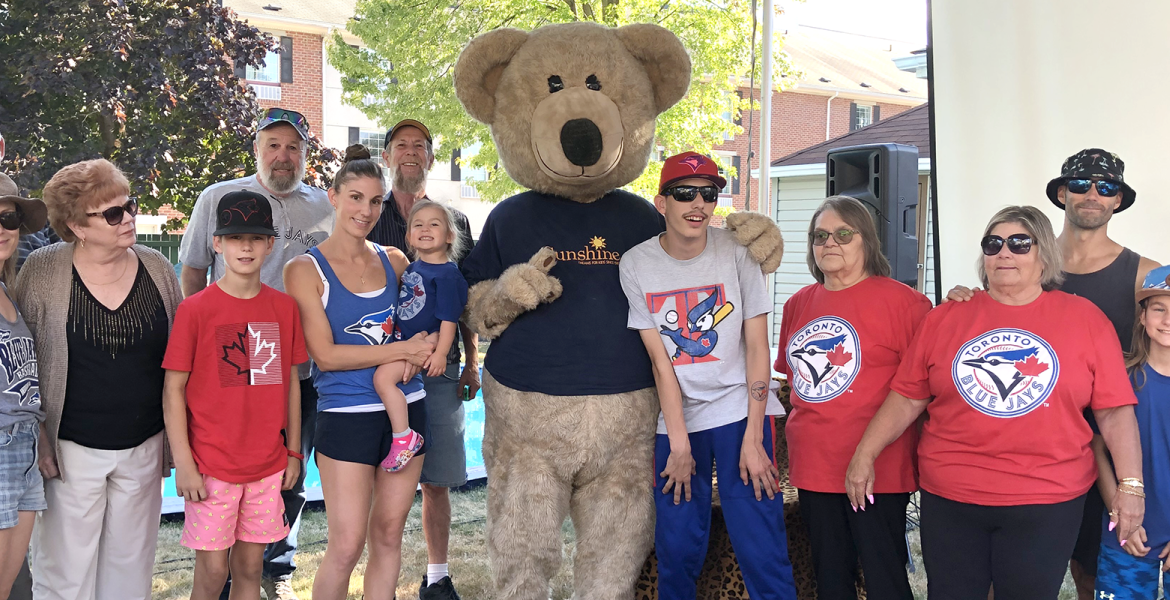 Family and friends surround young boy wearing sunglasses and blue jays t-shirt. Bear mascot stands beside him.