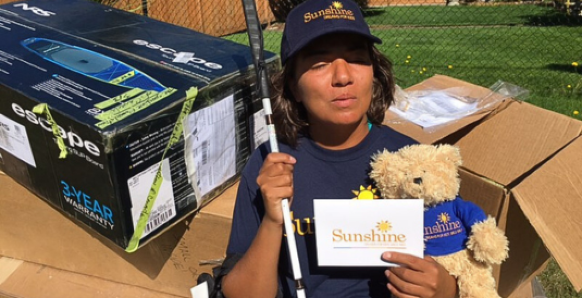 Veronica wears a Sunshine hat and is holding a teddy bear, Sunshine card, and her white cane. In the background is a box with her paddle board.
