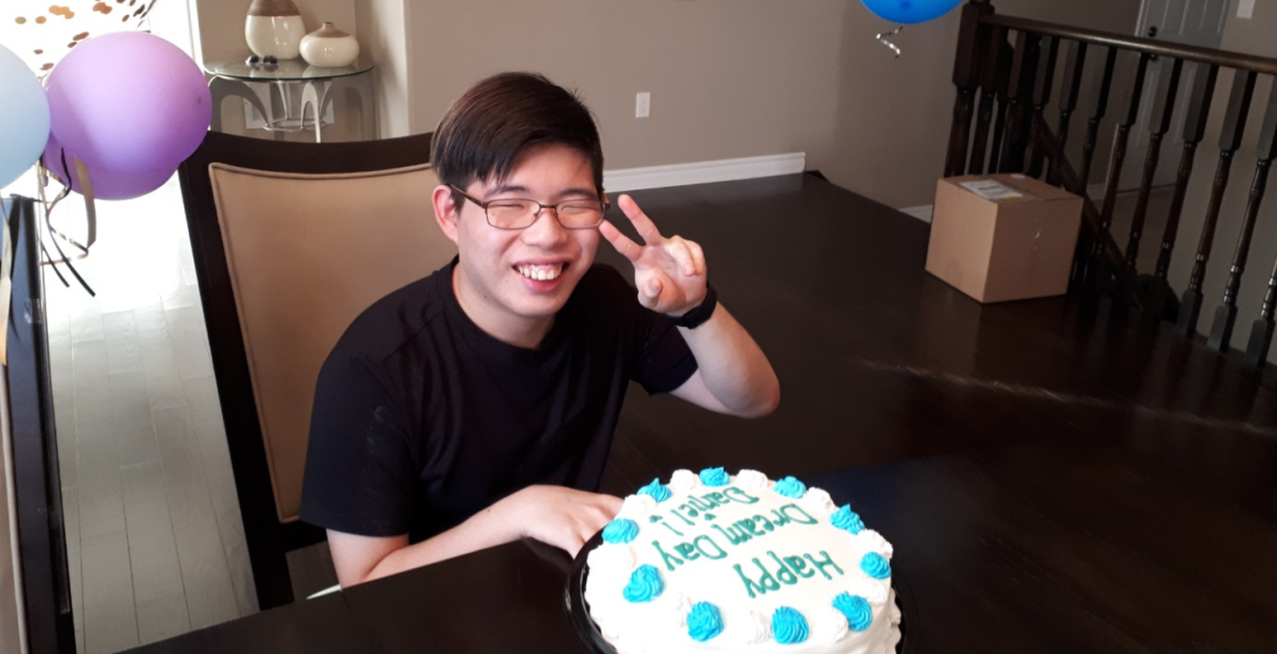 Young boy with glasses, asian, seated at his dining table in front of a cake, smiles with fingers up in peace sign