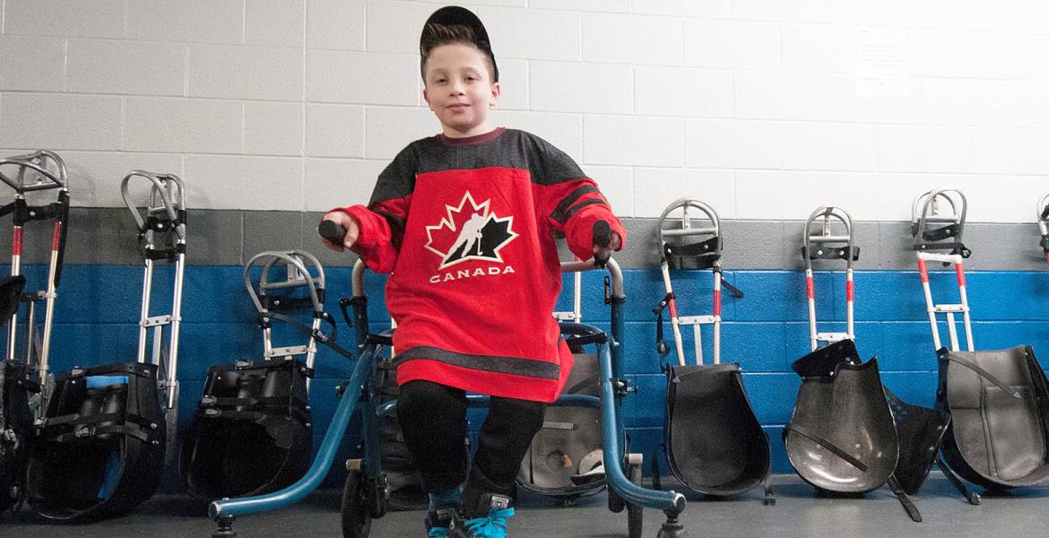 Young boy wearing baseball hat and red team canada jersey while leaning on his walker in a locker room full of sledge