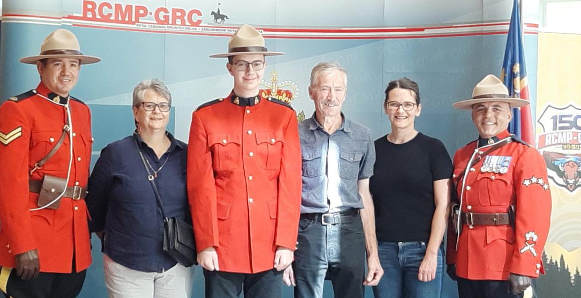 Erik stands in front of RCMP wall in uniform and hat, his parents on each side and two RCMP officers on each end