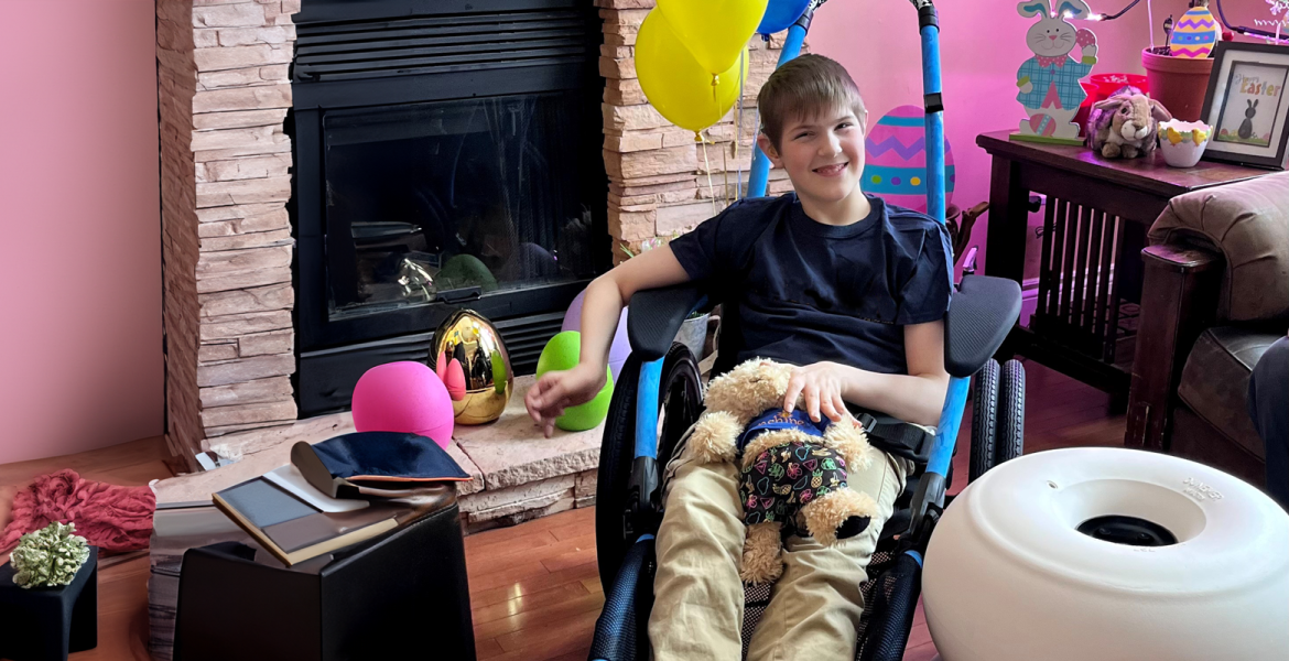 Boy seated in a blue hippocame chair with teddy bear on his lap and large white wheels