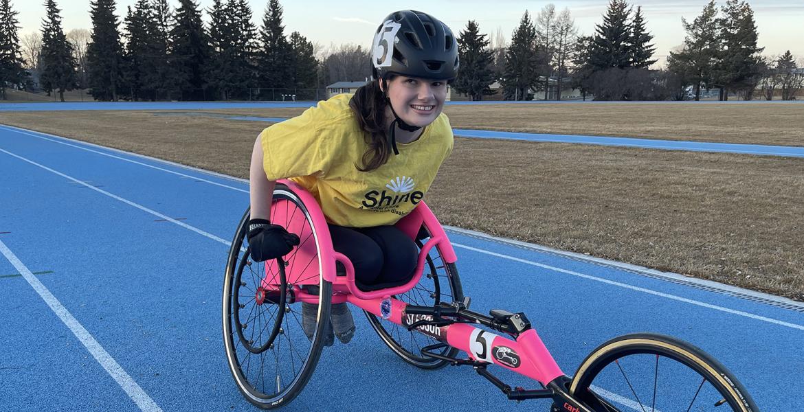 Maggie is seated in her hot pink racing wheelchair on a bright blue outdoor track wearing a yellow Shine tshirt and black helmet