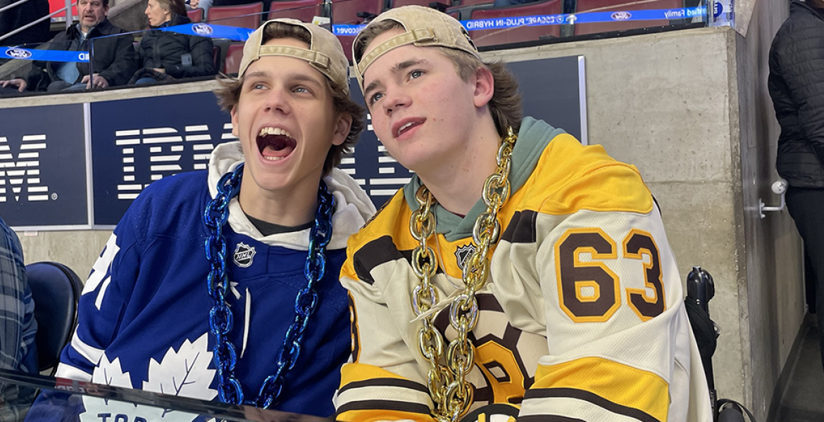 Young teen, seated with backwards baseball cap and Bruins jersey, beside his friend in a Toronto Maple Leafs jersey at a hockey game.