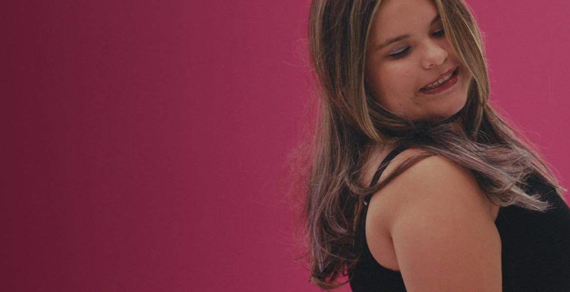 Scarlette, a blind indigenous teen, poses like a model with her eyes closed, looking over shoulder. Pink background