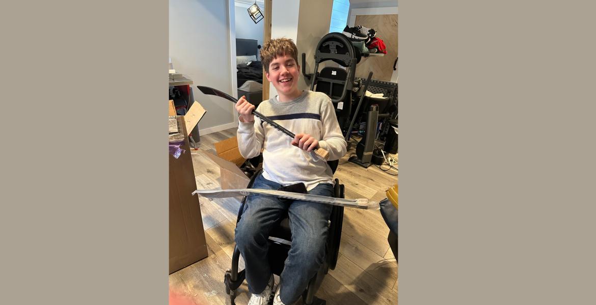 Blake seated in wheelchair holding sledge hockey sticks beside a large box