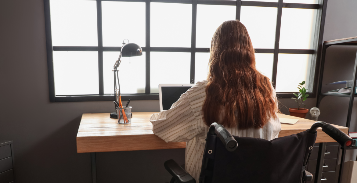 Female with long hair seated in wheelchair at a desk, view from behind her