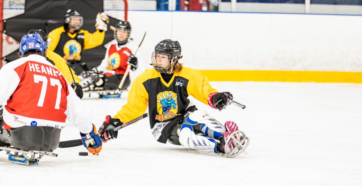 Girl in black and yellow hockey attire using an accessible hockey sledge, with her hockey stick trying to take a puck
