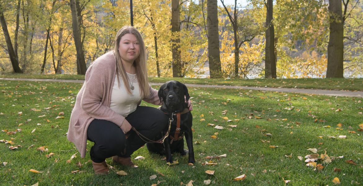 Kolby crouching beside her guide dog on the grass in a park