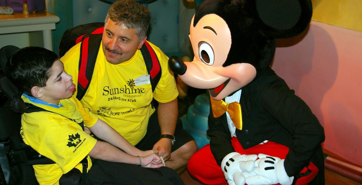 Marc in bright yellow t-shirt accompanies child in a wheelchair with matching t-shirt as they meet Mickey Mouse costume character.