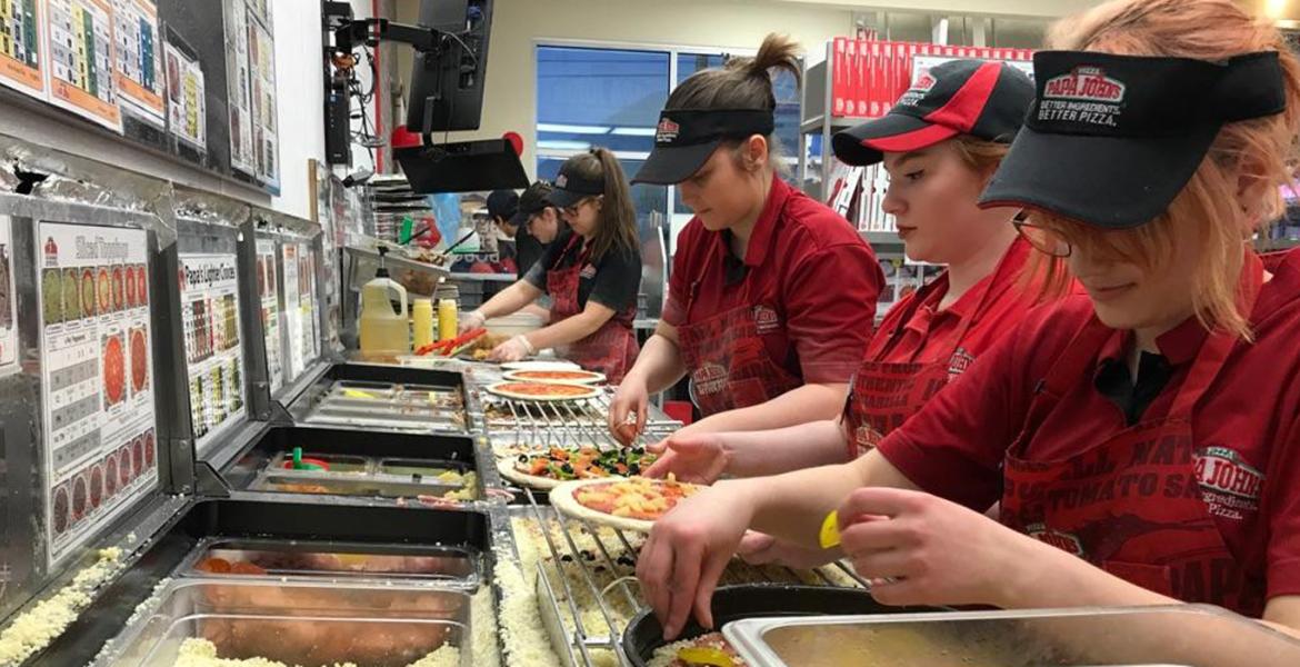 In the kitchen at Papa John's showing assembly line of workers topping pizzas