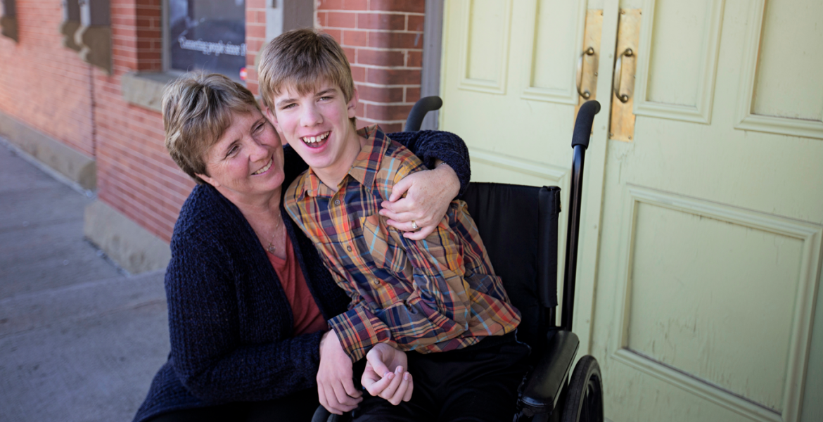 Young teenager with blonde hair sits in wheelchair, his mom embraces him from the side. They're in front of a brick building with large cream coloured doors