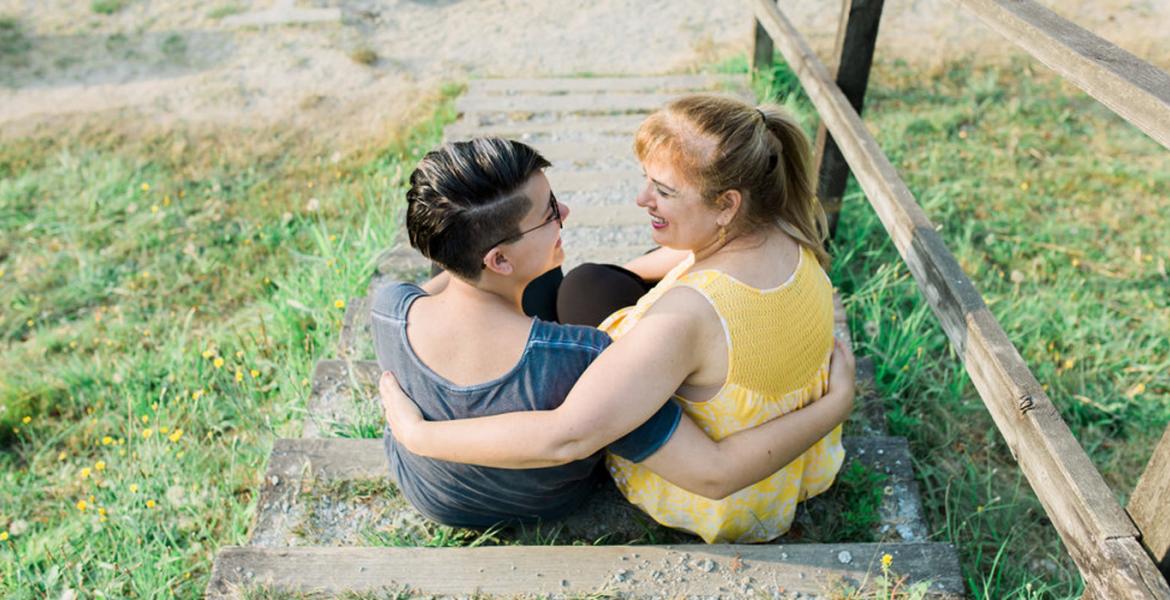 Kathy seated outdoors with back to us on cement stairs, embracing and smiling with partner