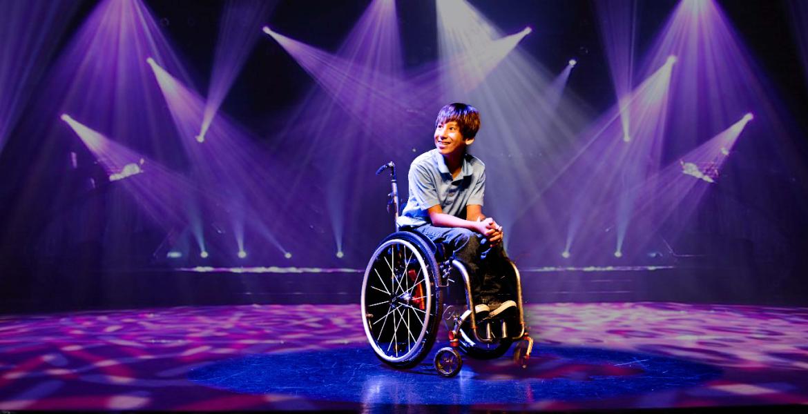 Tai seated centre stage in wheelchair surrounded by a backdrop purple spotlights