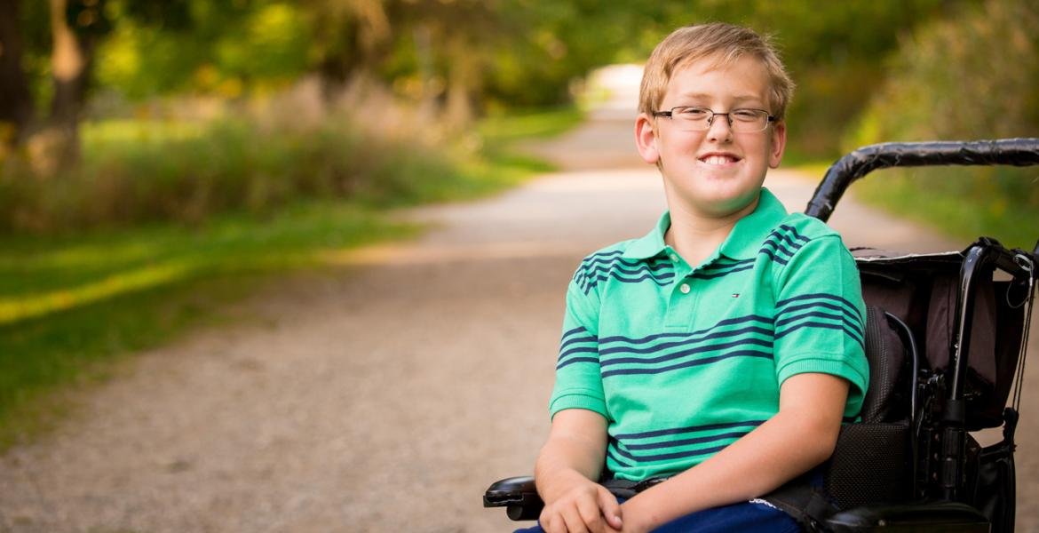 Child in wheelchair wearing bright blue striped polo smiling and surrounded by nature