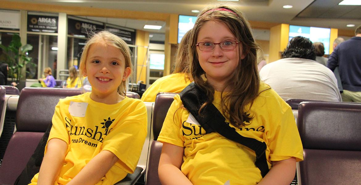 Two young girls smile in yellow Sunshine t-shirts while waiting at their gate at the airport