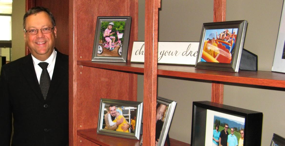 Wayne standing next to wooden shelving unit filled with framed photos