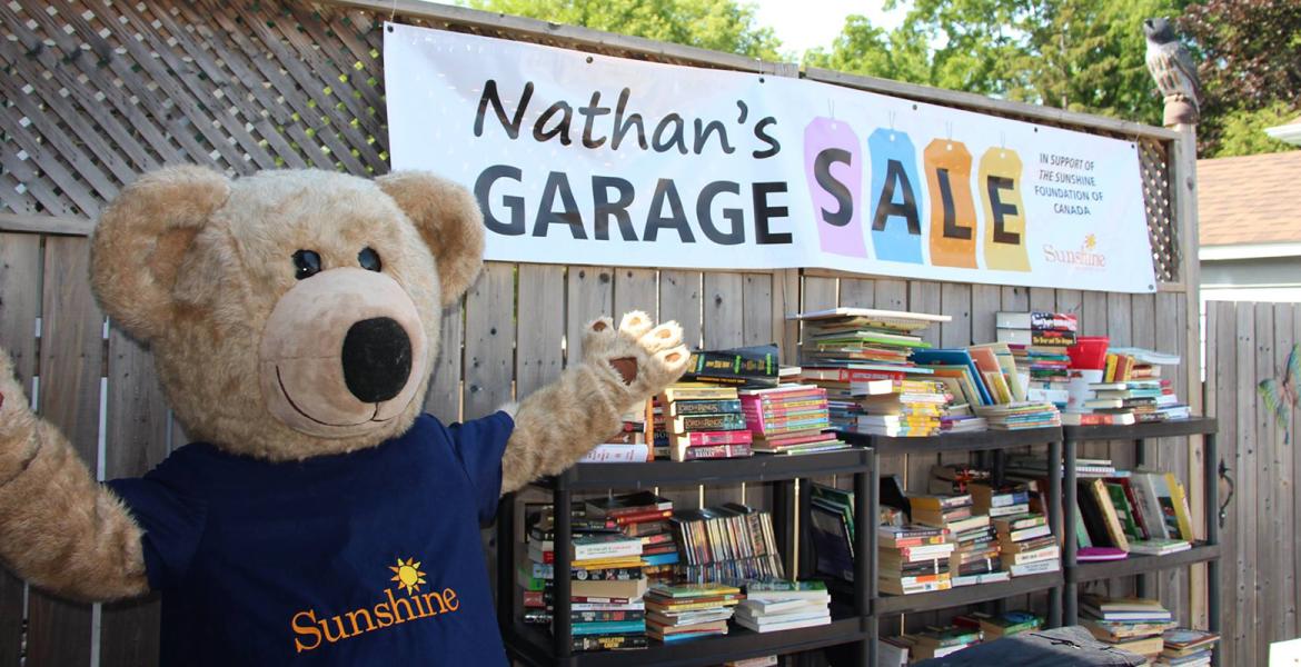 Large banner reading "Nathan's Garage Sale" hangs on a fence above a bookcase filled with books with large teddy bear standing next to the shelf